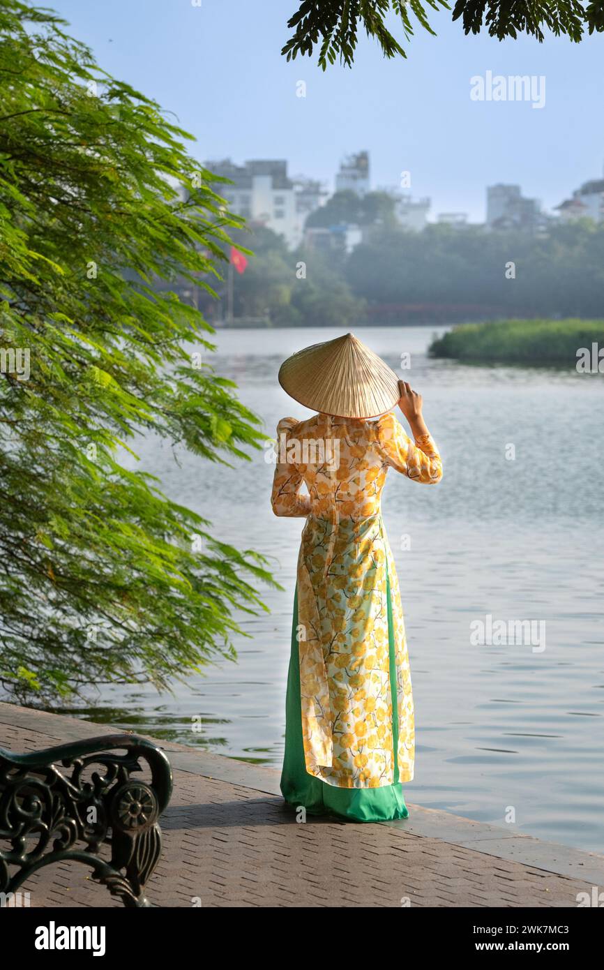 Woman in traditional dress and conical hat at Hoan Kiem Lake in Hanoi, Vietnam Stock Photo