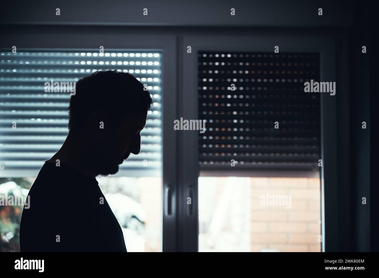 Depression and sadness, mental health concept. Silhouette of depressed hopeless male standing by the window with shutters pulled down. Selective focus Stock Photo