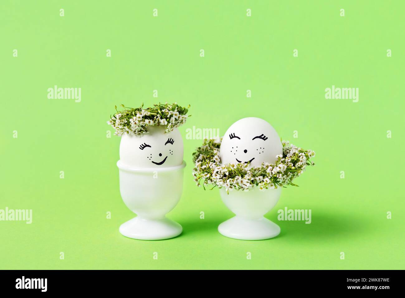 Two blue Easter eggs with funny face with spring flowers in white stands on a green background. Happy Easter concept. Greeting card. Stock Photo