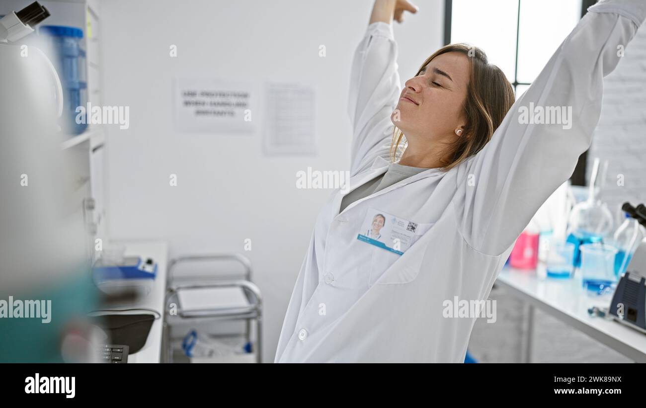 Tired yet determined, young attractive blonde woman scientist stretches arms while ambitiously working on computer in research lab Stock Photo