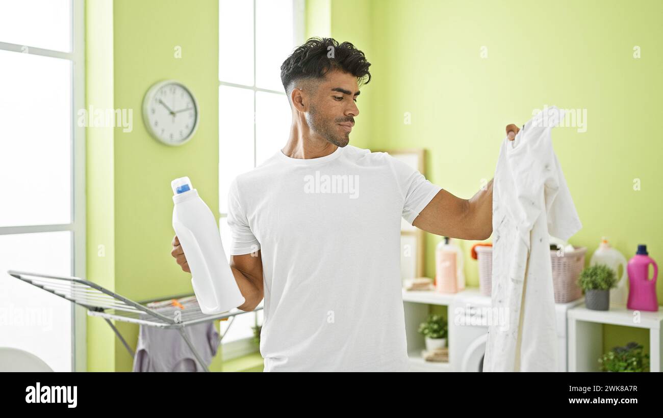 Handsome hispanic man doing laundry in a bright room, holding detergent and inspecting a white shirt. Stock Photo