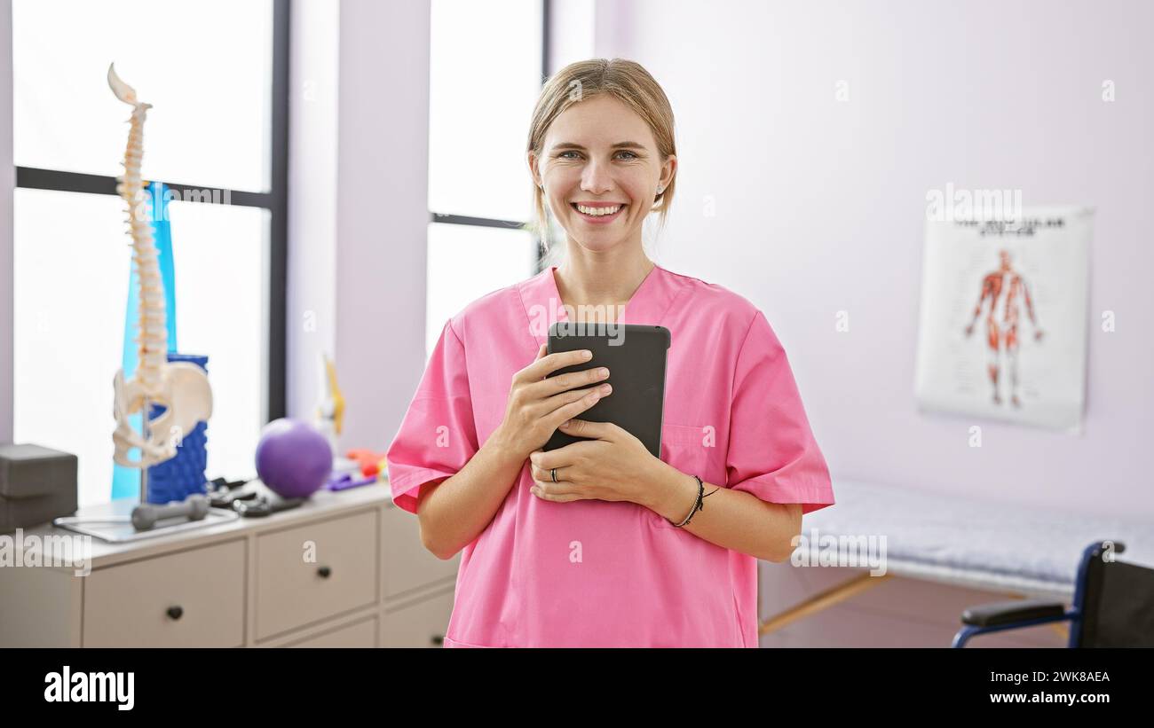 Smiling woman in pink scrubs holding tablet in a well-equipped rehabilitation clinic with anatomical posters Stock Photo