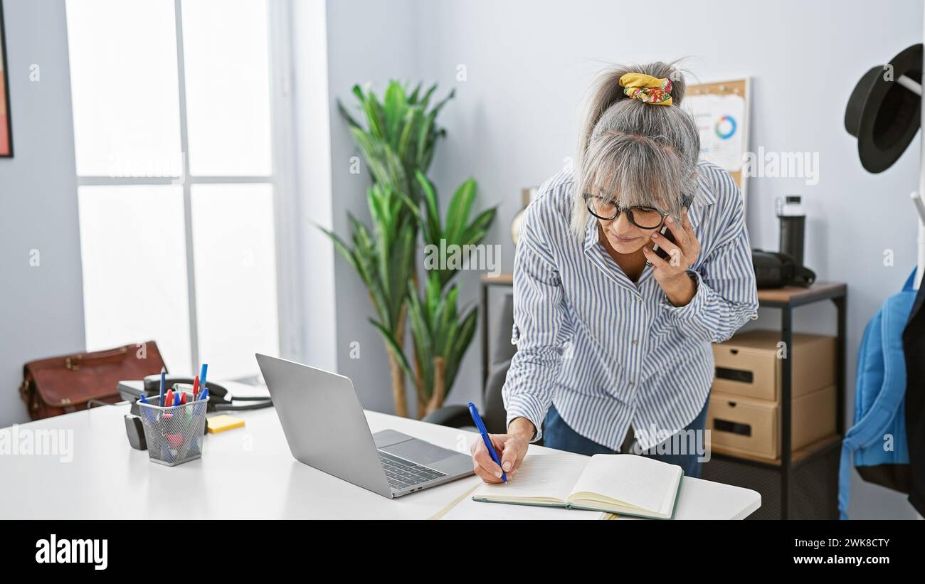 A mature woman multitasks in a modern office, talking on the phone ...