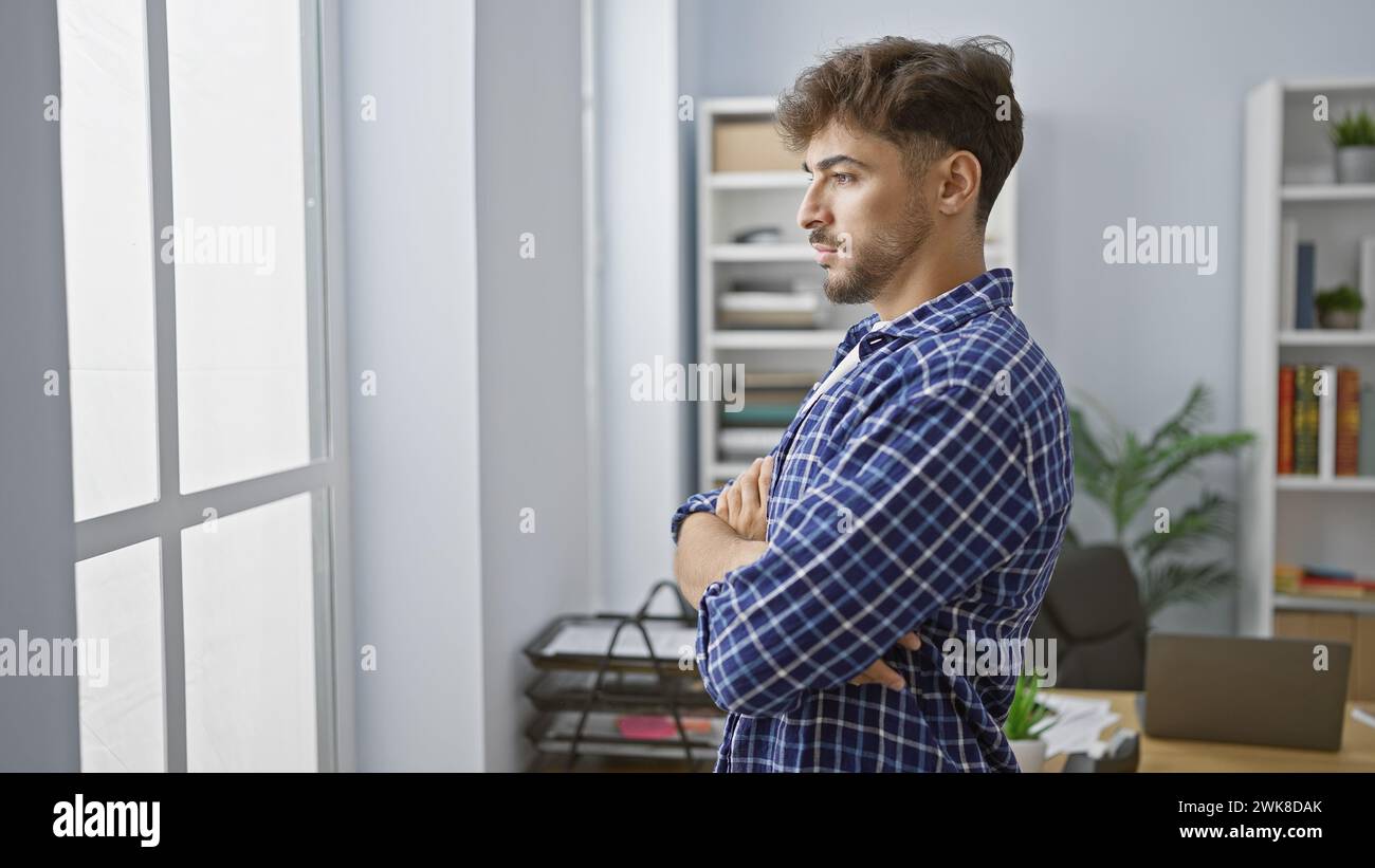Effortlessly elegant, young arab man doubtfully ponders, worker stands, arms crossed, gazing through office window. is he facing a problem, or brewing Stock Photo