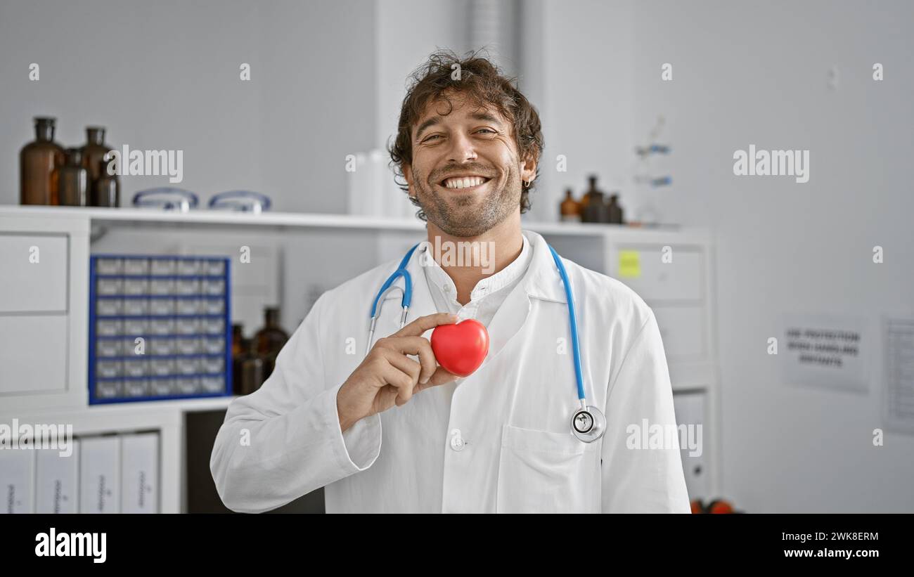 Smiling man in white lab coat holding a red heart in a hospital setting, conveying healthcare concept. Stock Photo