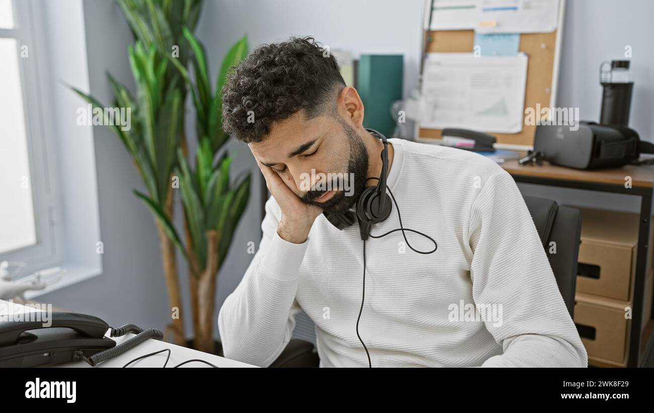 Tired hispanic man with beard using headset at office desk looking stressed Stock Photo