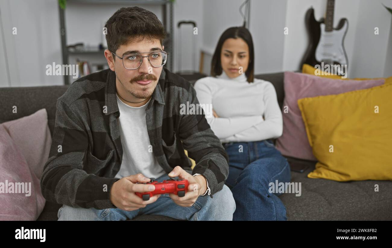 An upset woman with crossed arms sitting beside a focused man playing video games in a modern living room. Stock Photo
