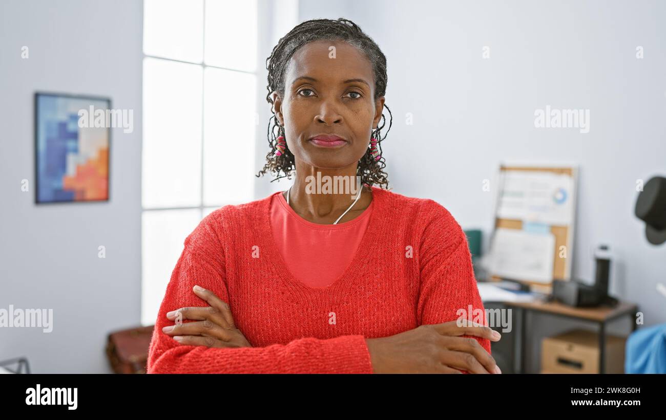 Confident african american woman in red sweater poses in a modern office setting, exemplifying professionalism and leadership. Stock Photo
