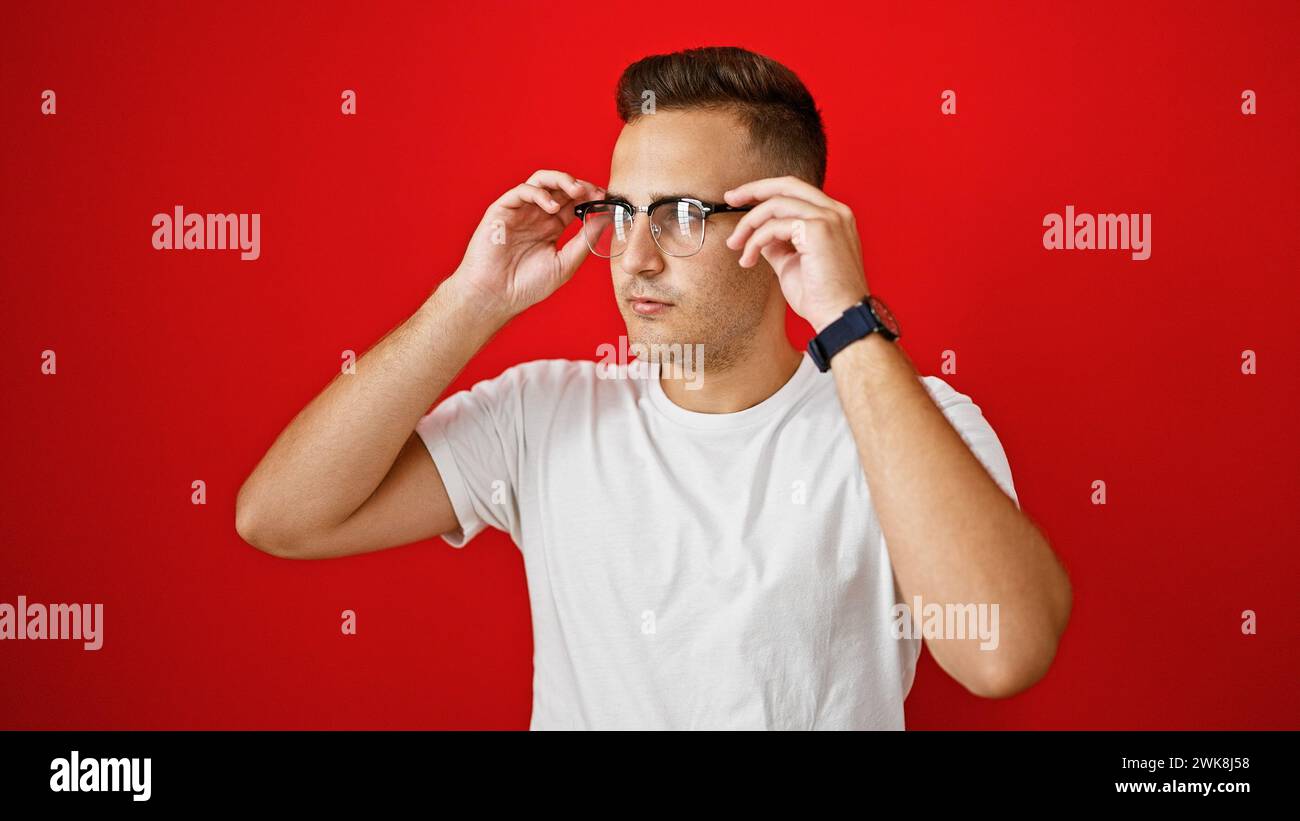 Handsome young hispanic man adjusting glasses against red isolated background, portraying confidence Stock Photo