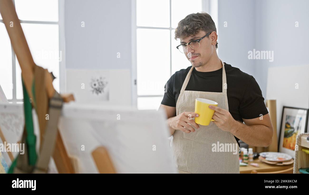 Determined young hispanic man in apron, an artist deep in thought, drinking coffee while looking at his drawing in art studio Stock Photo