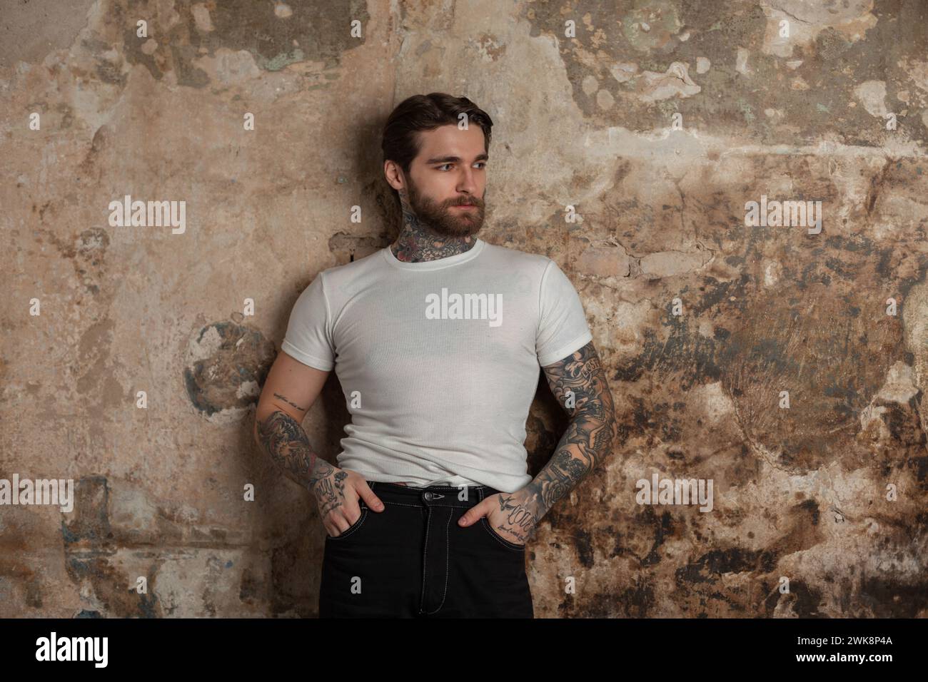 Cool Hipster Brutal Man With A Hairstyle And Beard In A Fashionable White Mock-up T-shirt With Tattoos Stands Near A Vintage Grunge Textured Wall Stock Photo
