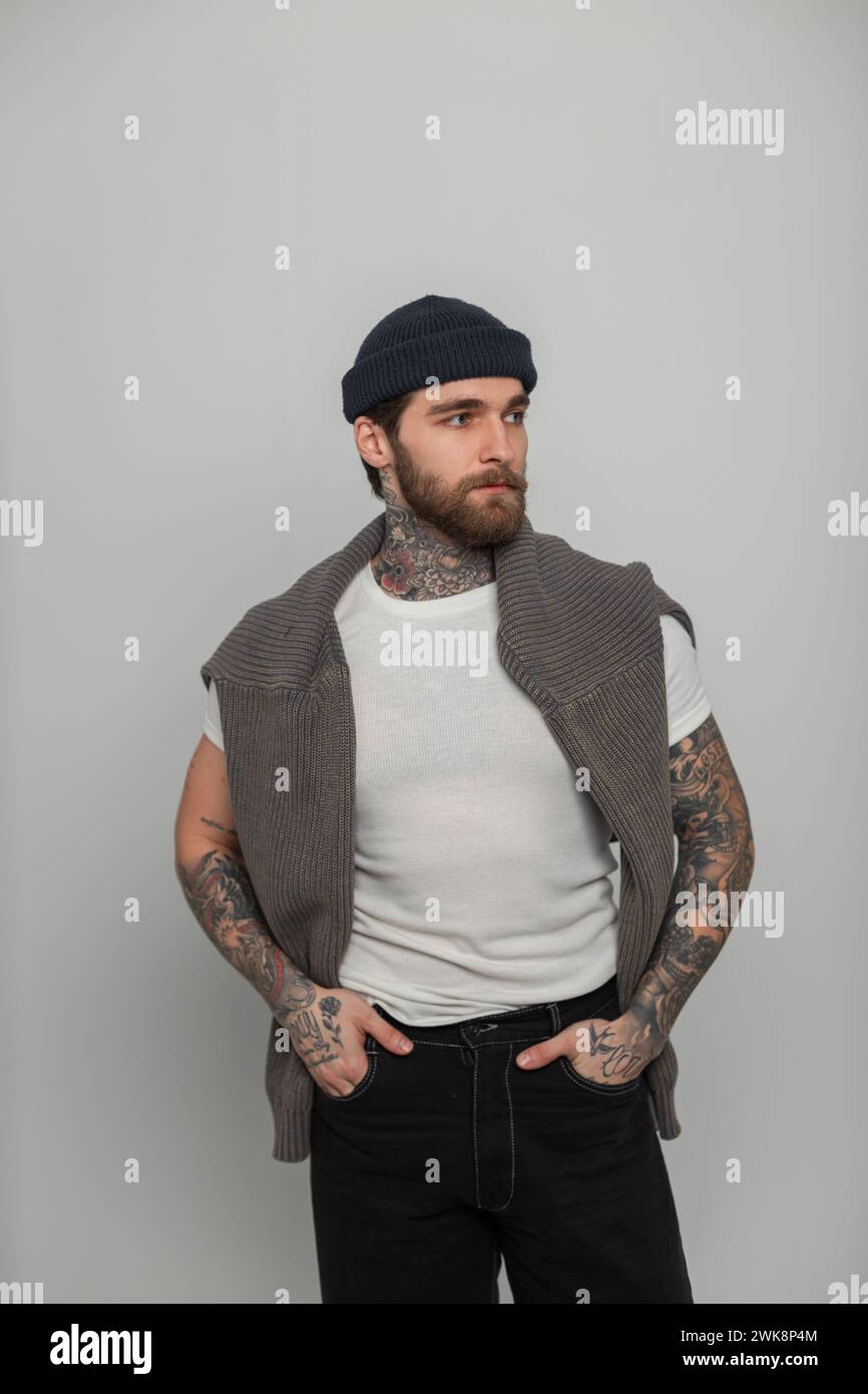 Handsome Fashionable Hipster Man With A Beard With Tattoos In A Fashion White T-shirt With A Knitted Sweater And Hat Stands On A White Background In T Stock Photo