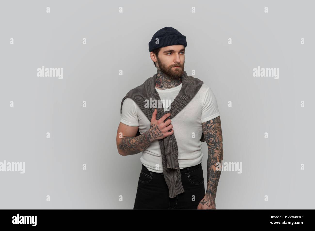 Cool Hipster Man With Tattoo With Hairstyle And Beard In A Fashionable White T-shirt And Knitted Sweater In The Studio Stock Photo