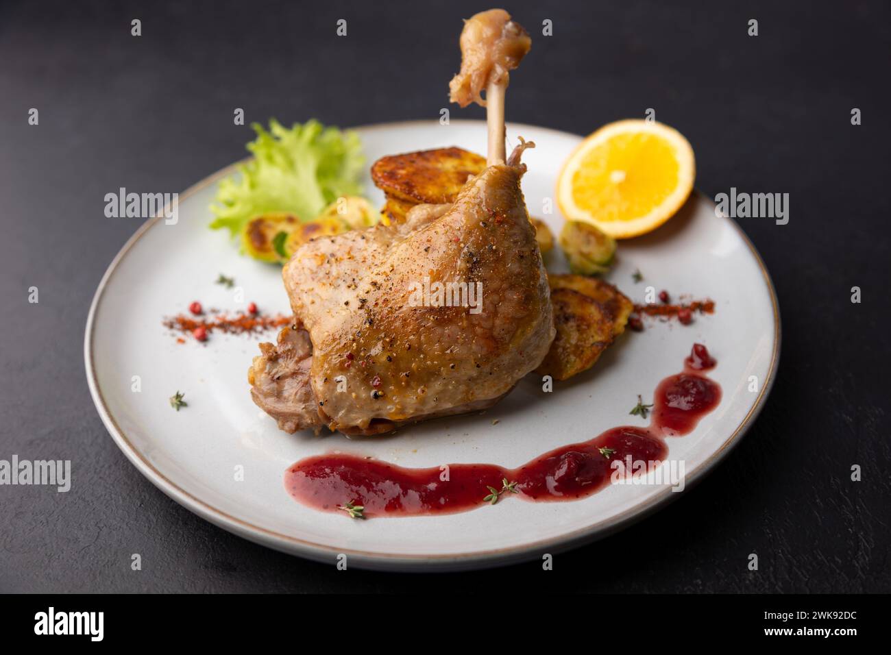 Duck leg confit with Brussels sprouts, baked potatoes, thyme, orange and lingonberry sauce. Traditional French cuisine. Selective focus, close-up. Stock Photo