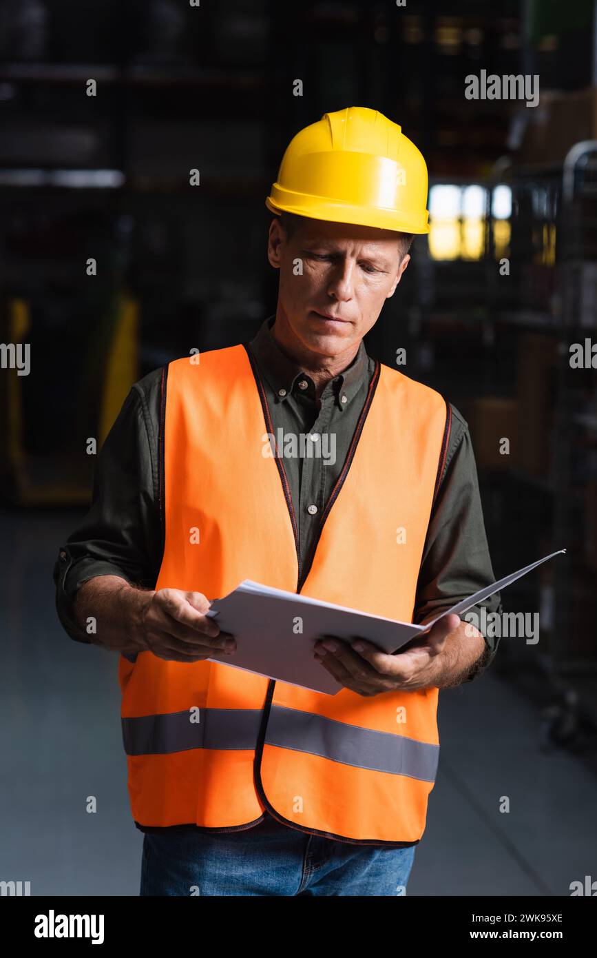 Focused middle aged supervisor in hard hat reviewing paperwork in warehouse, logistics and cargo Stock Photo