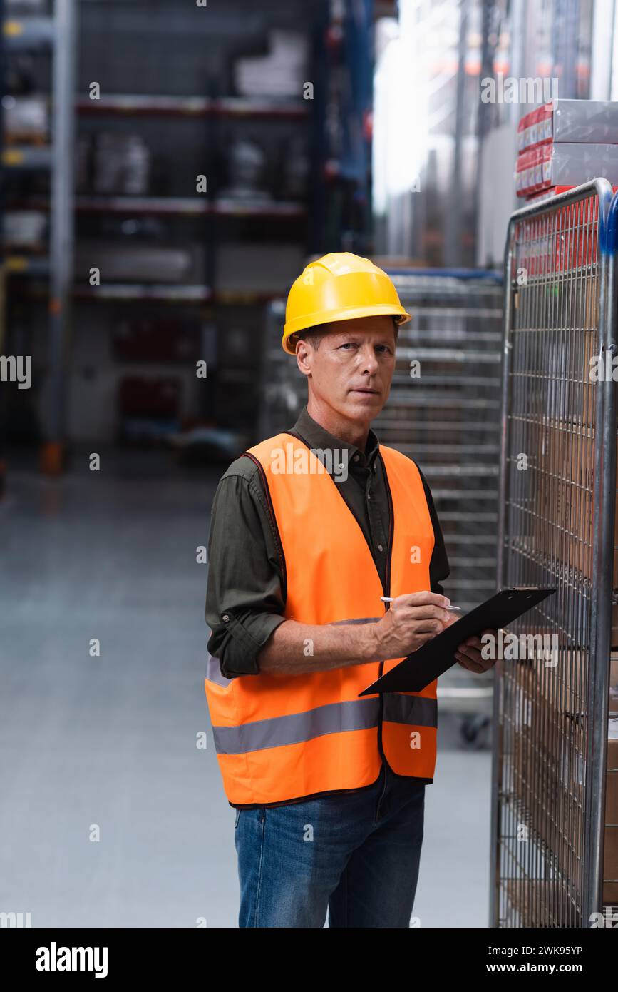 middle aged supervisor in hard hat and safety vest doing paperwork in warehouse, logistics and cargo Stock Photo