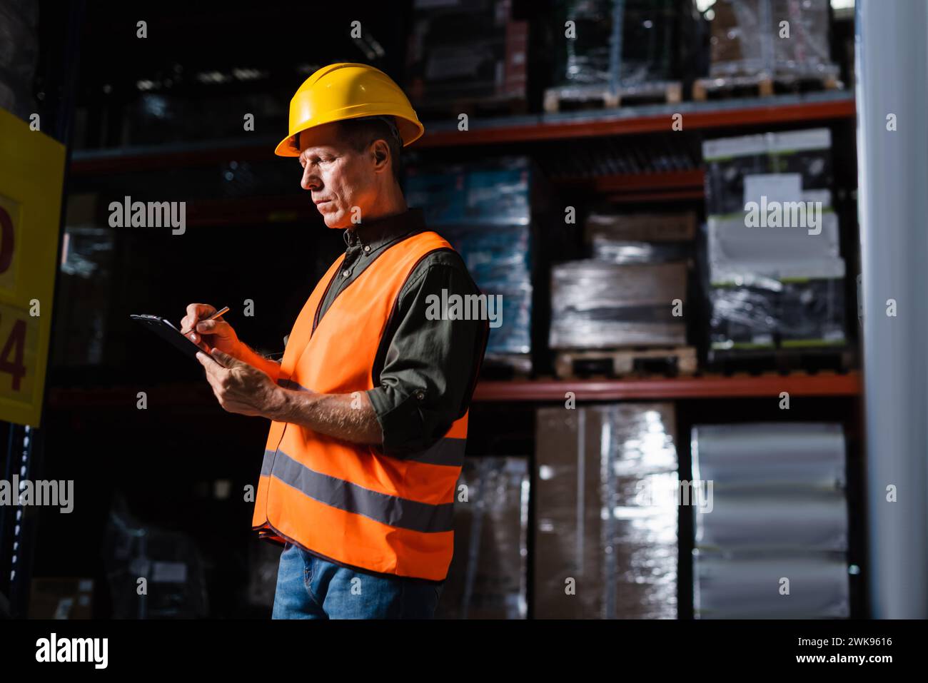 middle aged warehouse supervisor in hard hat taking notes on clipboard while inspecting cargo Stock Photo