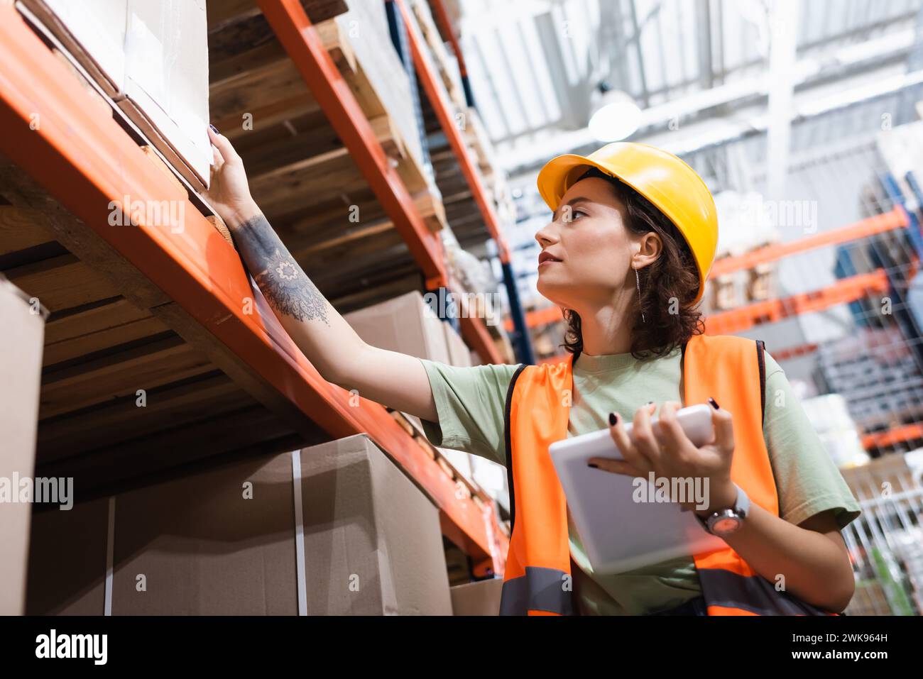 tattooed female warehouse worker in safety vest and hard hat holding digital tablet near cargo boxes Stock Photo