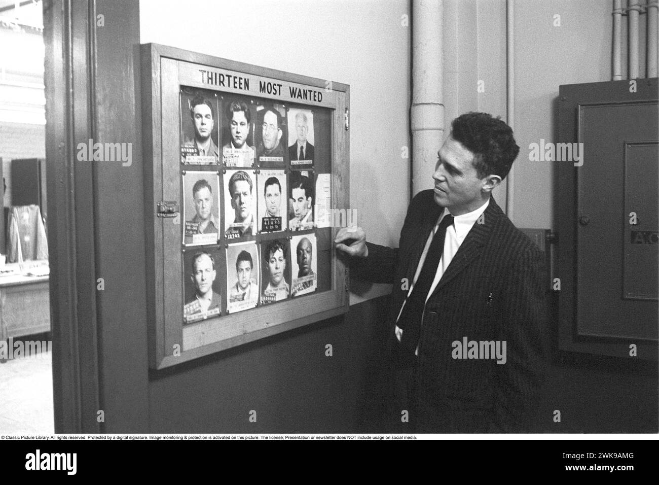 At a police station in New York USA in 1959. A detective by photographs on the wall with the heading Thirteen Most Wanted. They are portraits of men who have so far managed to avoid the police, but who in this way remind the police of what they look like.   Roland Palm ref DV11 *** Local Caption *** © Classic Picture Library. All rights reserved. Protected by a digital signature. Image monitoring & protection is activated on this picture. The license; Presentation or newsletter does NOT include usage on social media. Stock Photo