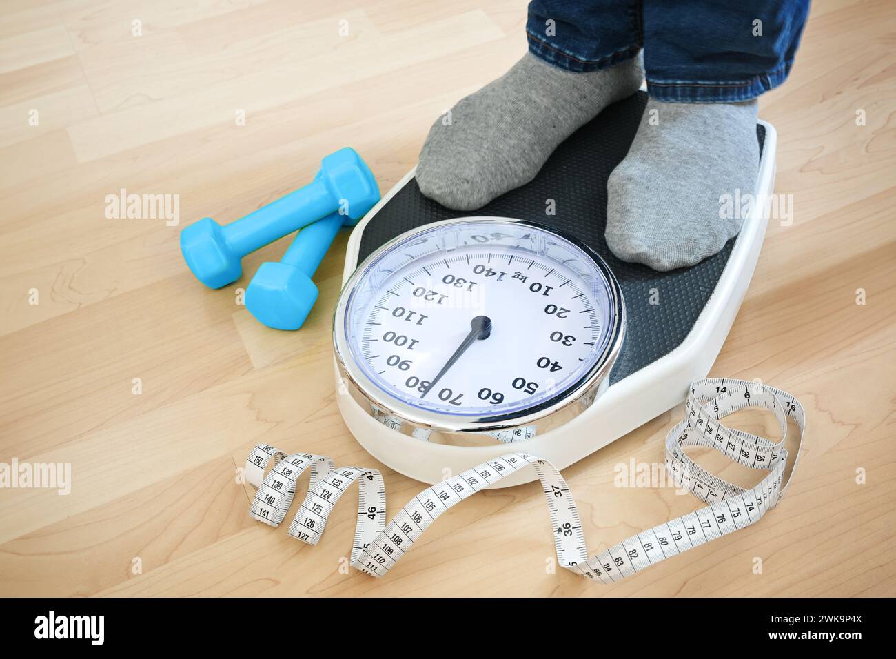 Feet of a man in gray socks standing on a scale to check the weight after fitness training, blue dumbbells and a measuring tape lying nearby on a wood Stock Photo