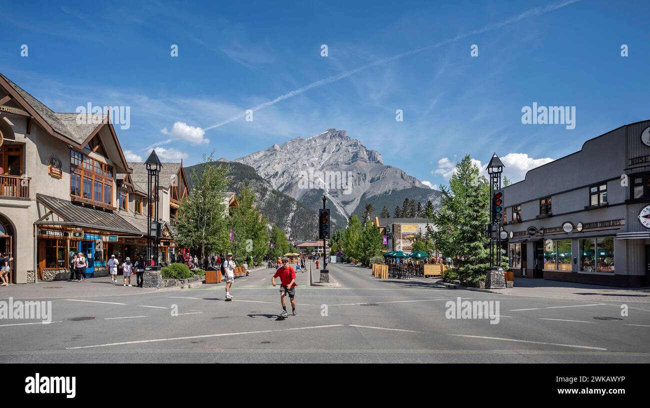 Skate boarders on Banff Avenue with snow capped Cascade Mountain in background in Banff, Alberta, Canada on 8 June 2023 Stock Photo