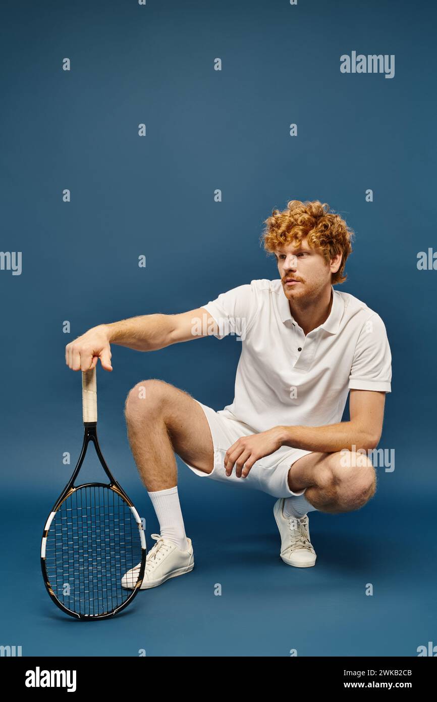 trendy redhead man in white tennis outfit sitting on haunches with racquet on blue, old money style Stock Photo