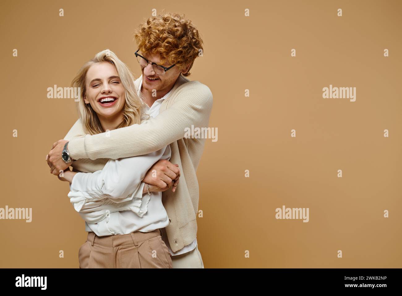 smiling redhead man in eyeglasses hugging blonde stylish woman laughing on beige, old money style Stock Photo