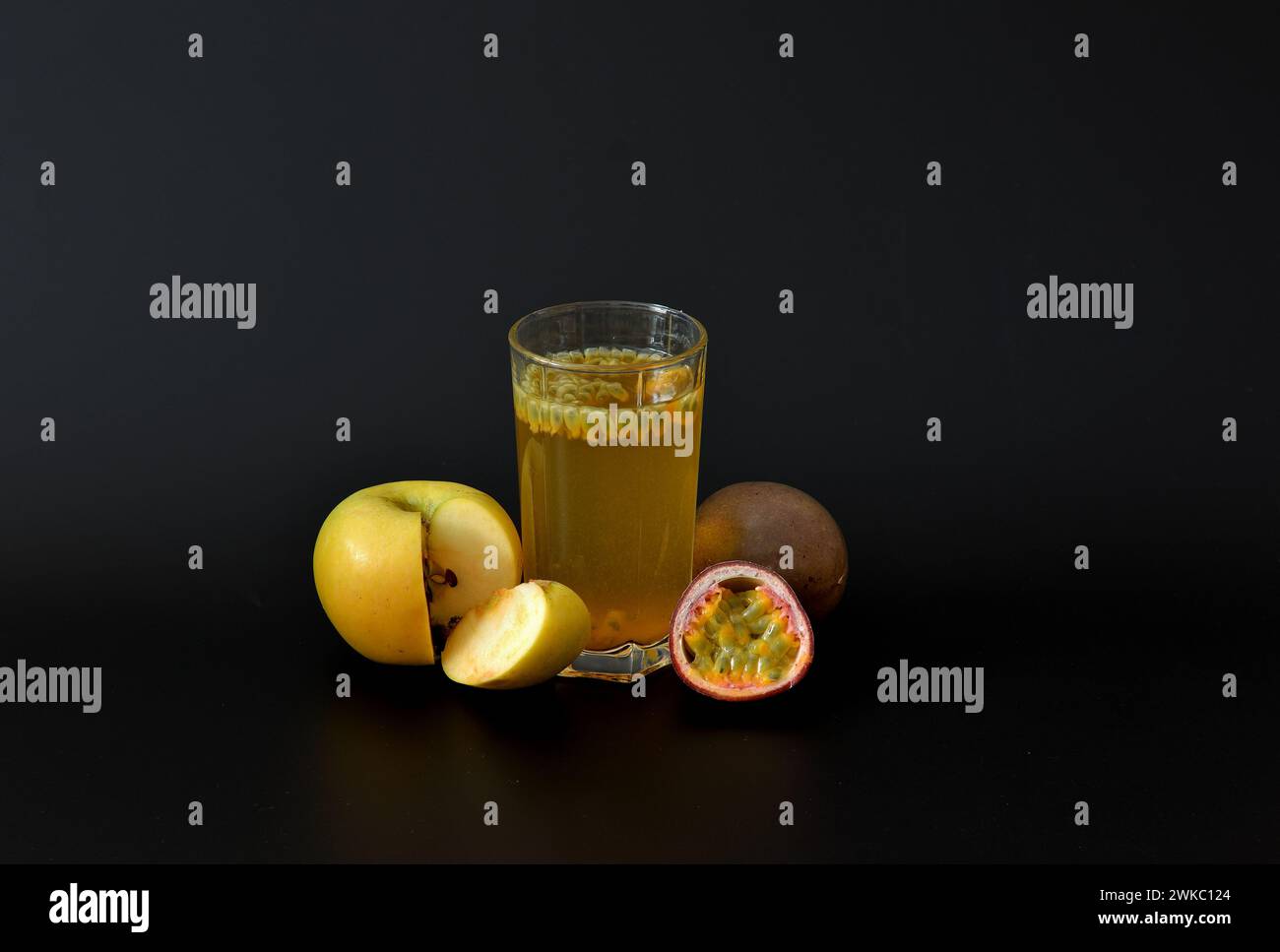 A glass of fresh fruit juice with seeds on a black background, next to a ripe yellow apple and pieces of passion fruit. Close-up. Stock Photo