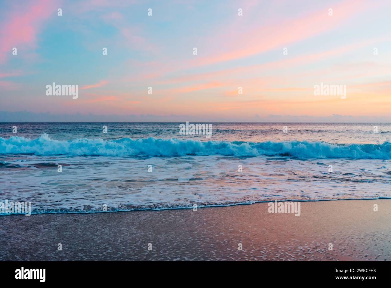 The sun sets over the tranquil beach, casting warm hues across the sky and reflecting its pink and golden glow on the gently lapping waves Stock Photo