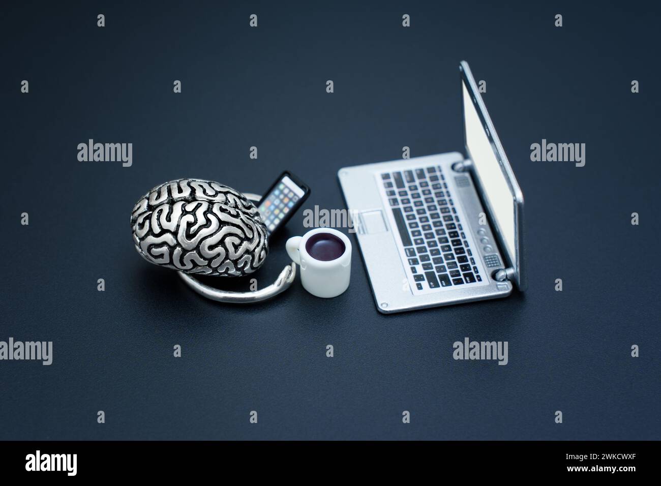 Miniature setup showcasing a steel brain model with coffee cup and cell phone working at laptop isolated on black. Stock Photo