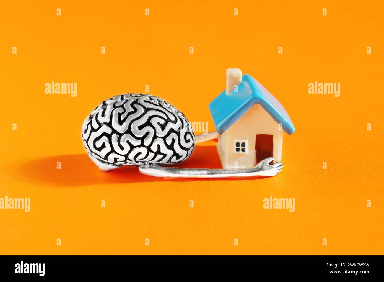 Steel human brain model tenderly cradling a miniature home in its hands against an orange backdrop. Inner world and mental well-being related concept. Stock Photo