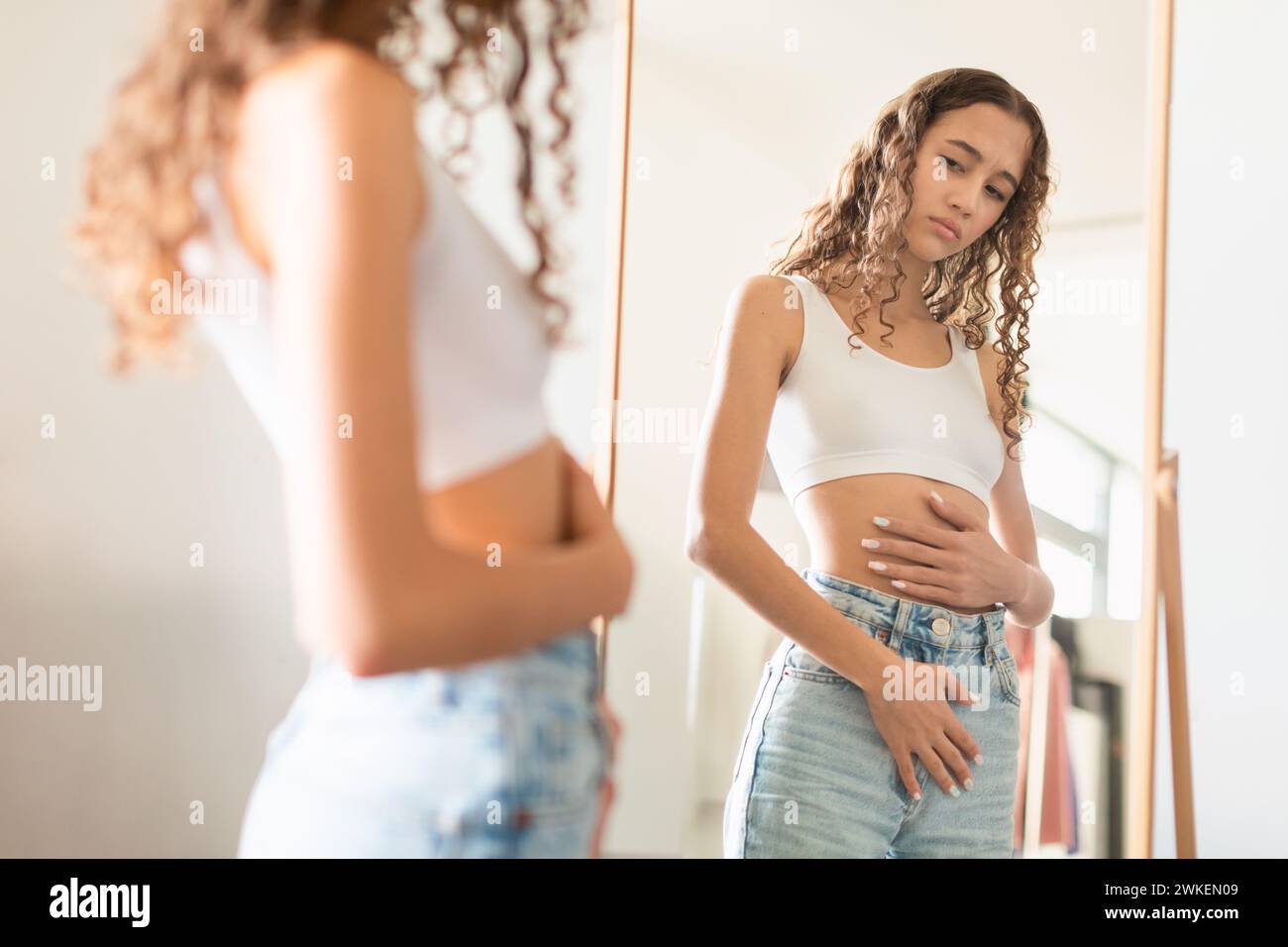 Unhappy teenage girl touching stomach in front of mirror indoor Stock Photo