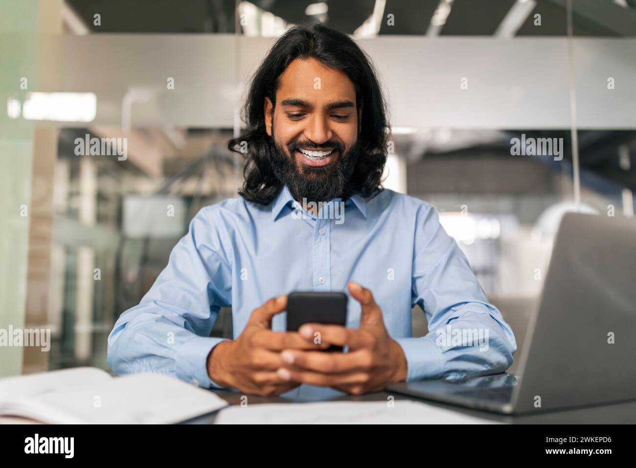 Positive indian guy manager using smartphone, modern office interior Stock Photo