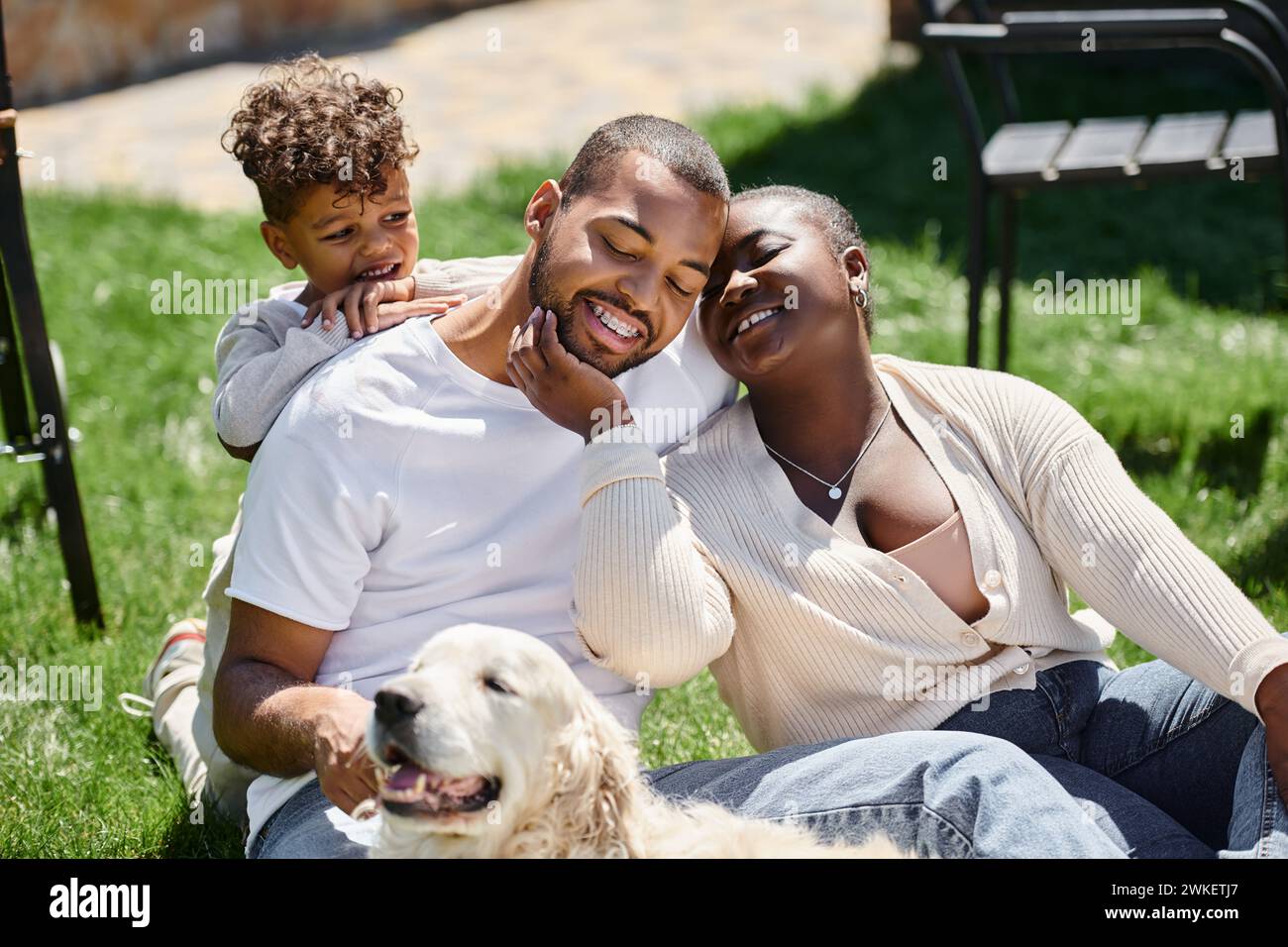 family moment of happy african american parents and son smiling and sitting on green lawn near dog Stock Photo