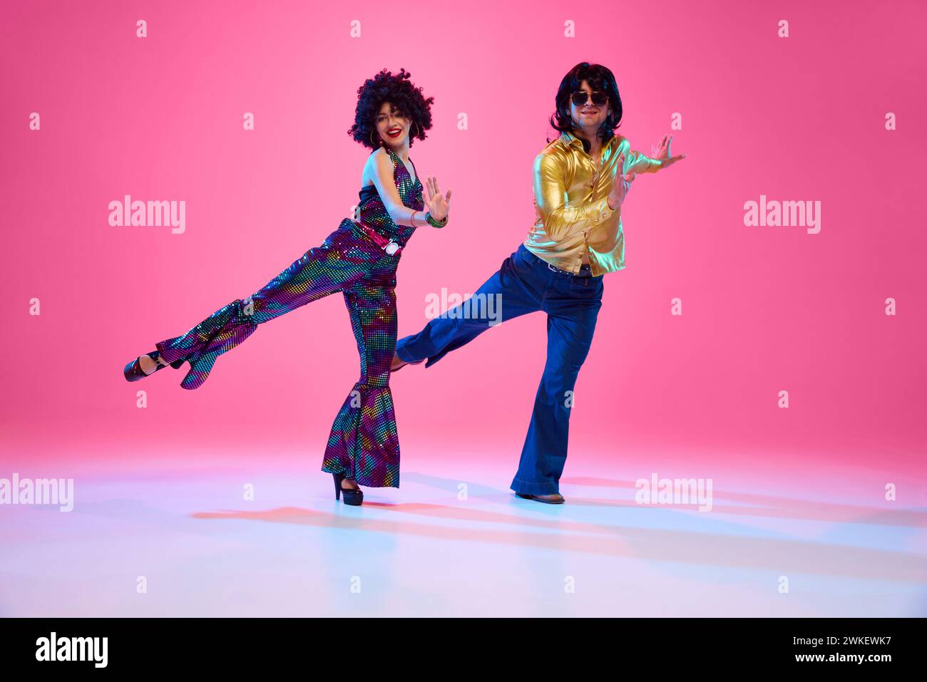 Retro disco era fashion. Couple with funky hairstyles and groovy clothes dancing energetic dance against gradient pink studio background. Stock Photo