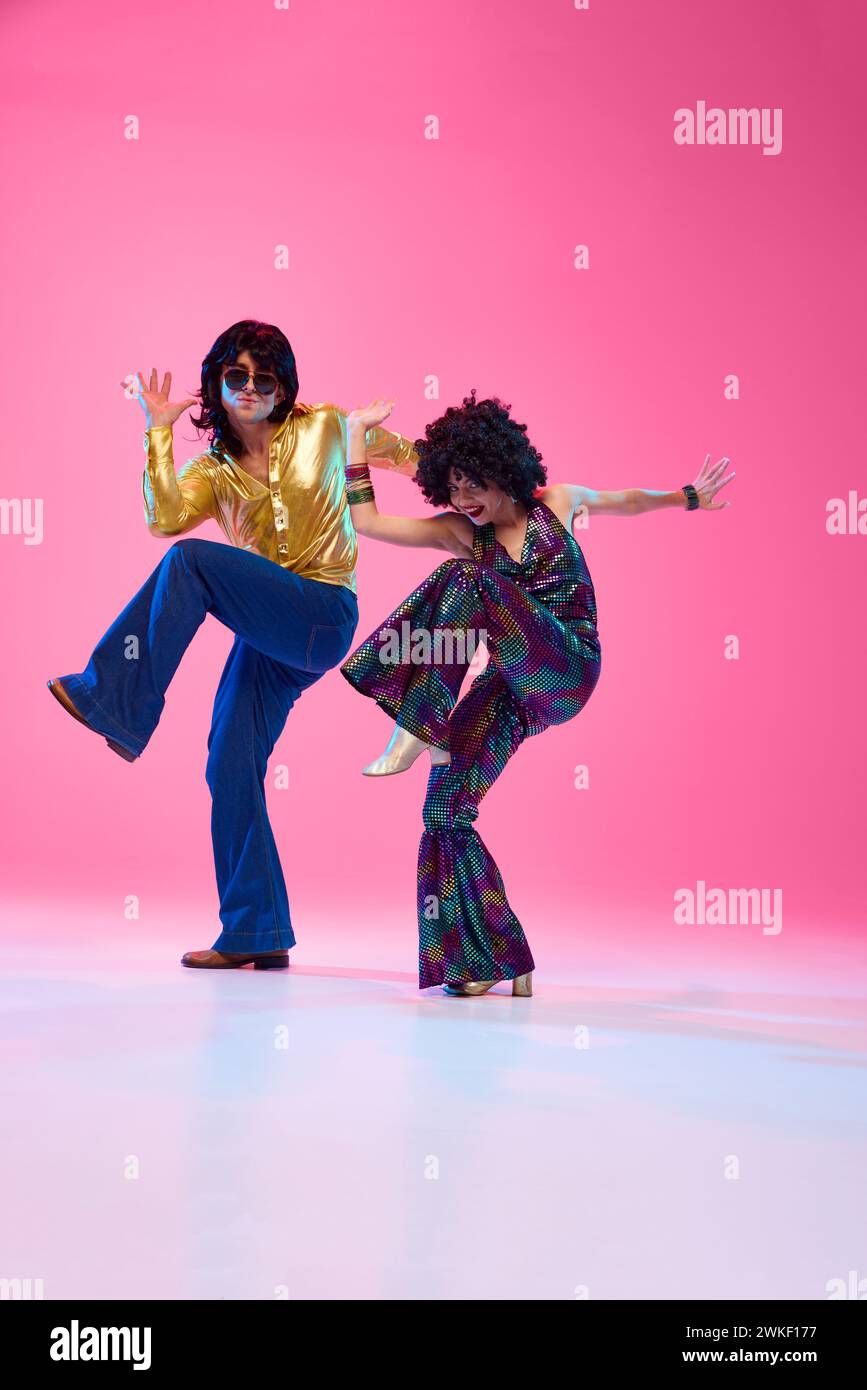 Enthusiastic disco dancers, man and woman in colorful 70s attire dancing in motion against gradient pink studio background. Stock Photo