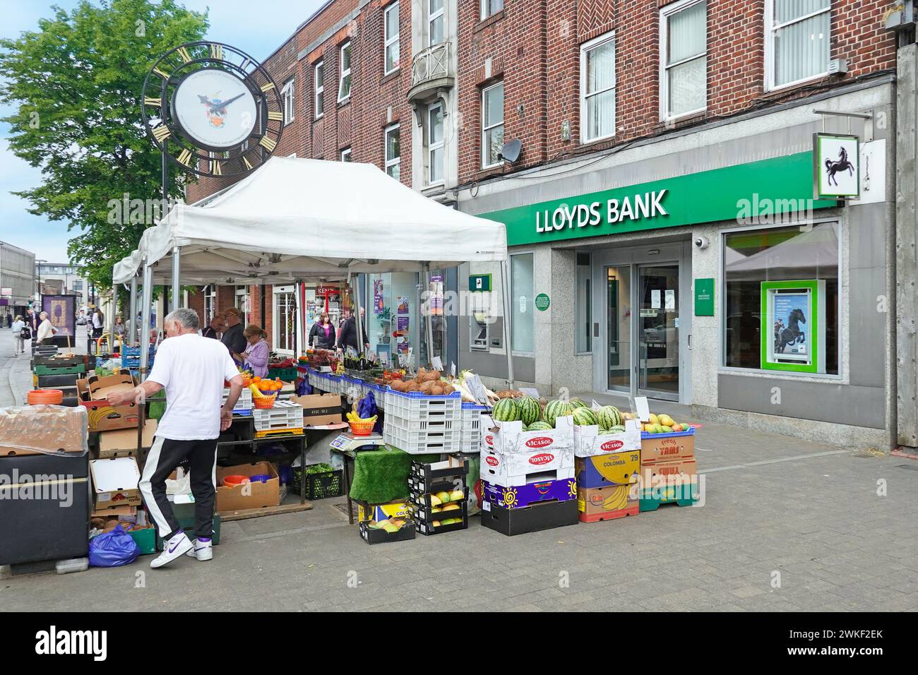 Market stall trader working his fruit & vegetable licensed stall pitch on pavement outside Lloyds Bank below town clock High Street Brentwood Essex UK Stock Photo