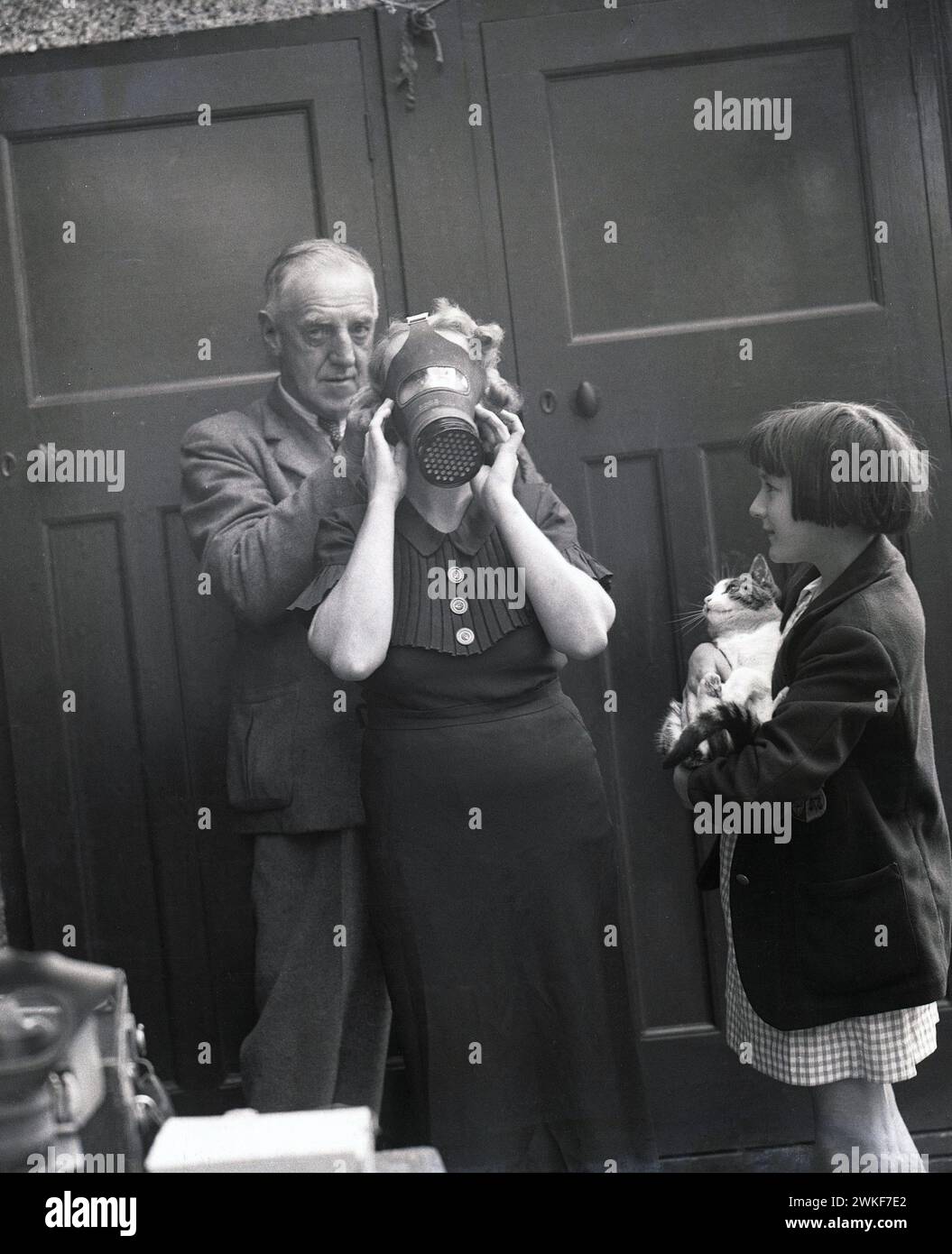 1940s, historical, wartime, a schoolgirl, holding pet cat, watching her grandmother try on a gasmask, England, UK. In the late 1930s, with war with Germany on the horizon, the Britsh government issued gas masks for the public at large, as protection against the dropping of posioned gas bombs, as memories were still strong of the horrors of gas attacks duting WW1. Stock Photo