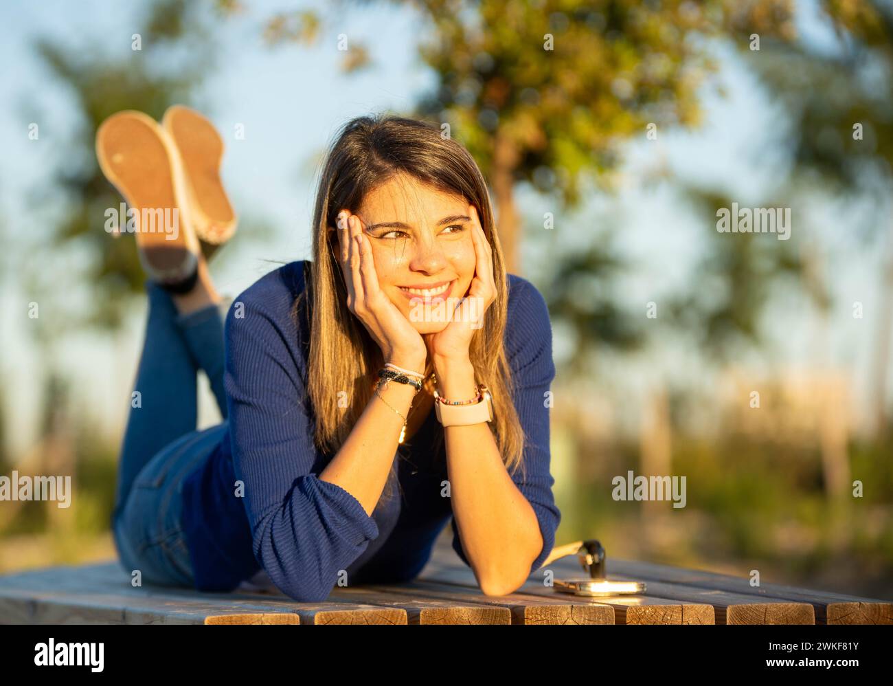 Happy and relaxed woman smiling while lying on a bench outdoors in a park during the sunset. Stock Photo