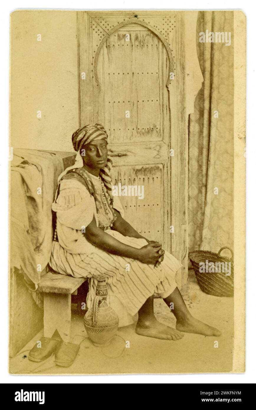 Original Victorian era CDV  Carte de Visite ( visiting card or CDV) albumen photograph of beautiful young servant woman in traditional Moroccon dress, sitting on a bench with ornate wooden door behind, looks like a studio set up, Tangier, Morocco. 1870's Stock Photo