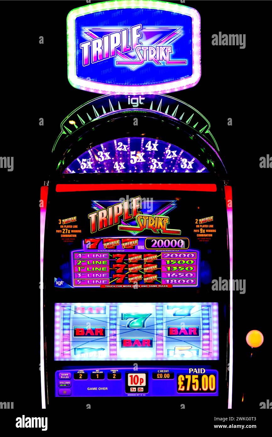 IGT Triple Strike video slot machine with illuminated belly and lozenge header panel graphics attracts players inside an amusement arcade. Stock Photo