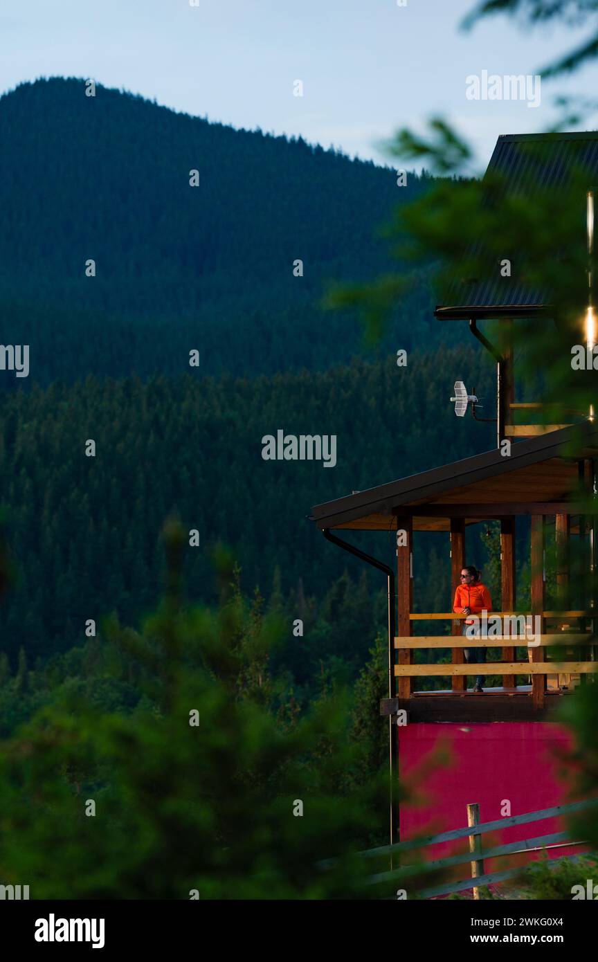 a girl in an orange jacket stands on the terrace of a wooden house against the background of mountains, a house for tourists in the Carpathians of Ukr Stock Photo