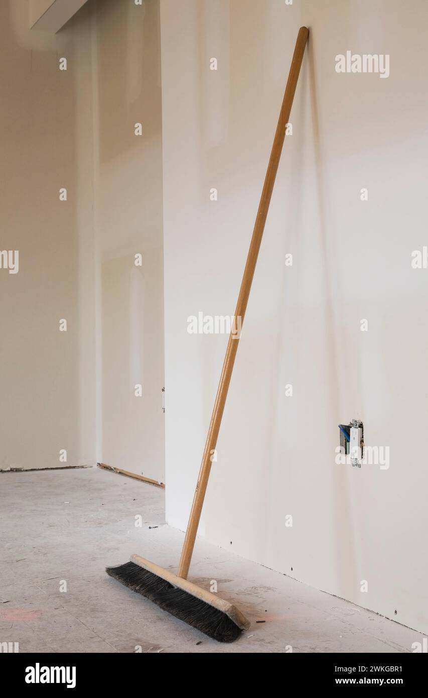 Sweeping broom leaning against unpainted plasterboard wall in unfinished room with dusty plywood floor inside home. Stock Photo