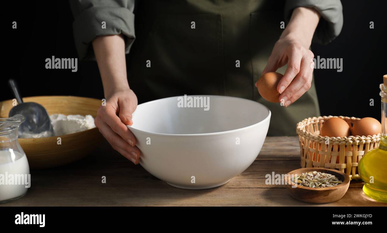 Making bread. Woman adding egg into dough at wooden table on dark