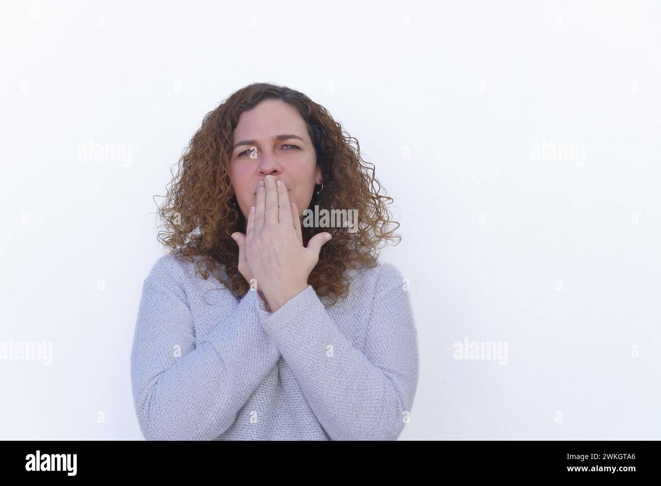Attractive blonde woman with curly hair and blue eyes covering her mouth with her hands looking at the camera with white background and copy space Stock Photo