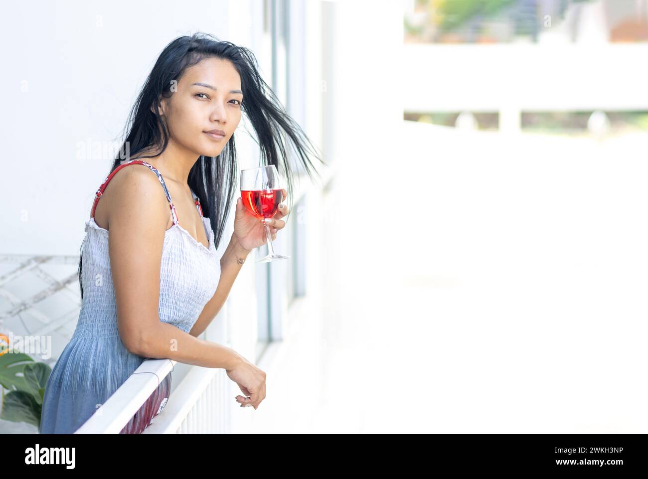 A woman in a summer dress holds a full wine glass in the balcony of a residential building Stock Photo