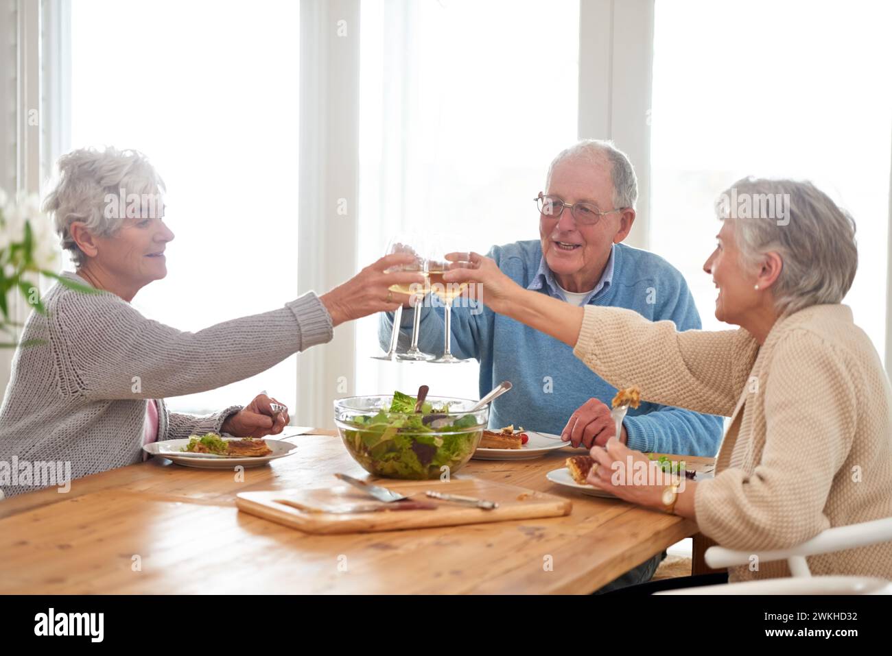 Wine, cheers and senior friends at brunch in home with smile, celebration and bonding in retirement. Food, drinks and toast with glass, old man and Stock Photo