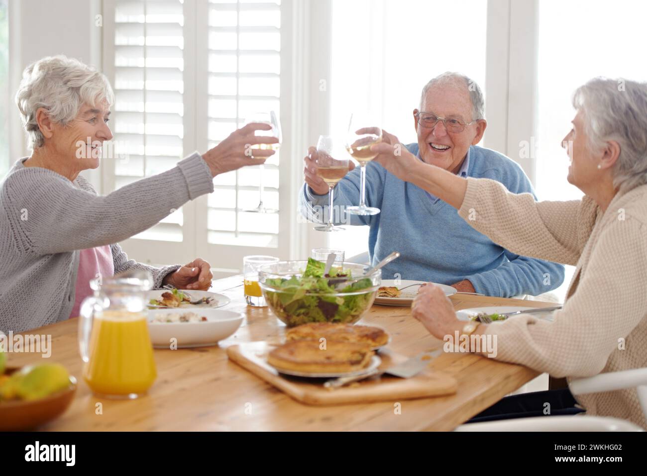 Wine, cheers and senior friends at lunch in home with smile, celebration and bonding in retirement. Food, drinks and toast with glass, old man and Stock Photo