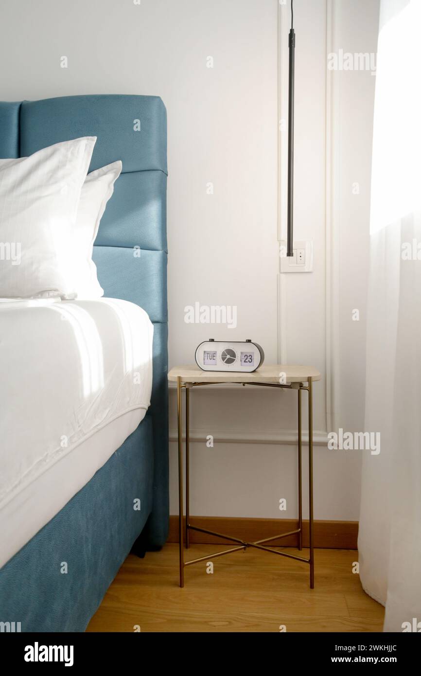Blue king size bed with crisp white bedding and digital alarm clock on the nightstand under beautiful morning sunlights Stock Photo