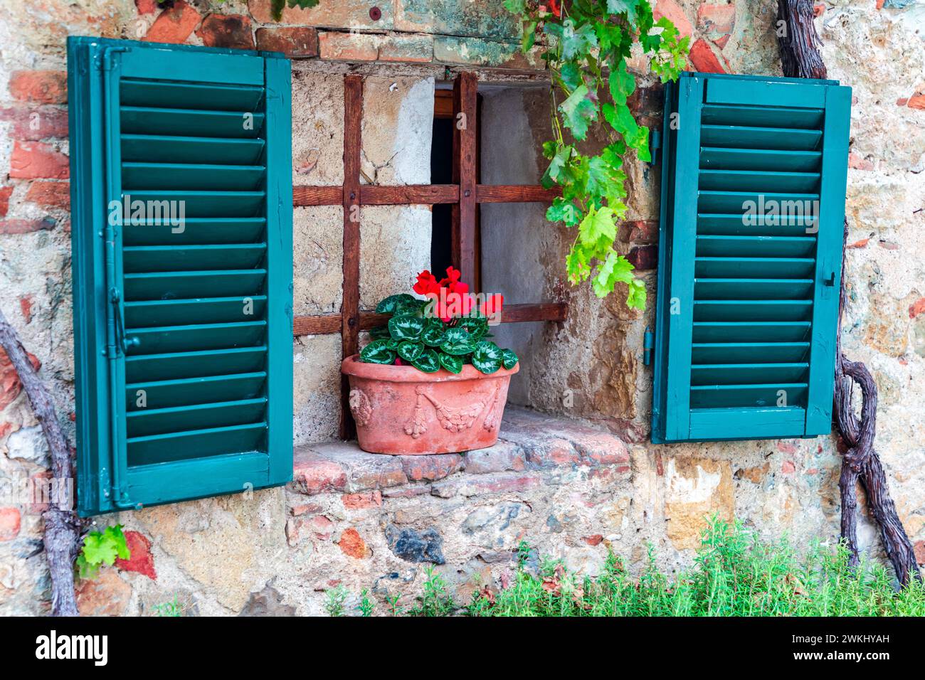 Small fairy tale style window in old stoned house with open green shutters. Clay made flower pot with red flower on stoned windowsill. Monteriggioni, Stock Photo
